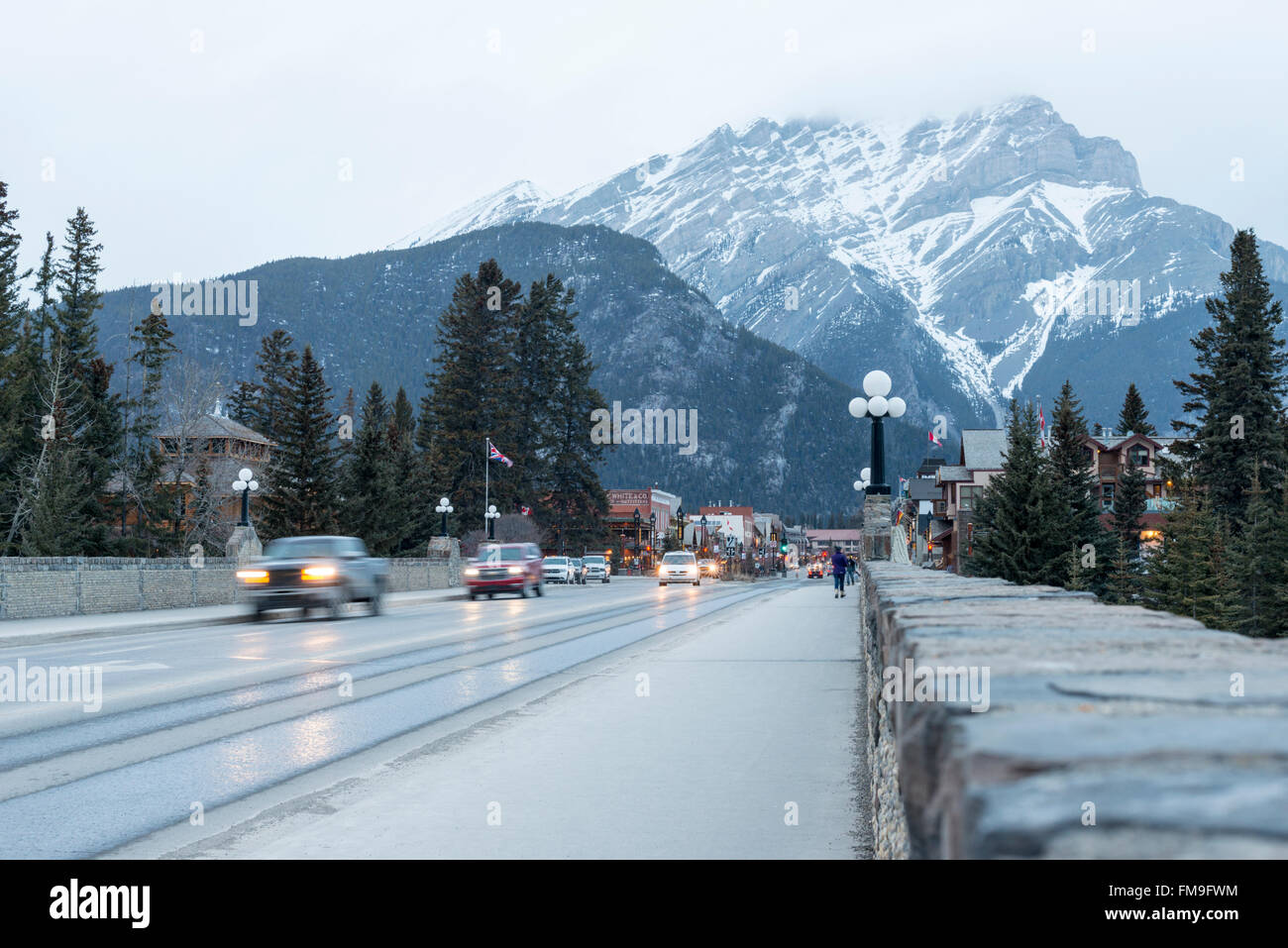 Traffic on Banff Avenue on the bridge over the Bow river at dusk Banff Canada with the Rocky mountains in the background Stock Photo