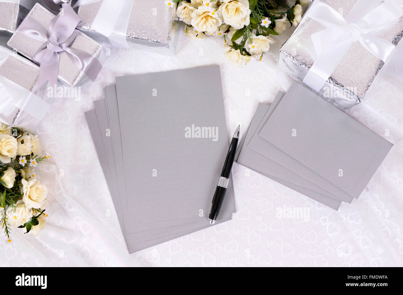 Silver gray writing paper or invitations with envelopes laid on bridal lace with several wedding gifts and white rose bouquets. Stock Photo