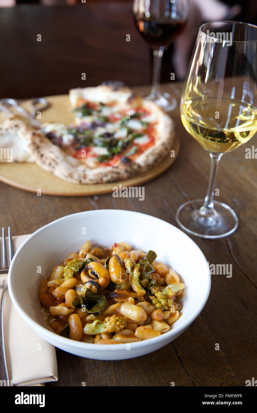 A bowl of white bean, romanesco and mussels soup with white wine and a pizza on the table behind it. Stock Photo