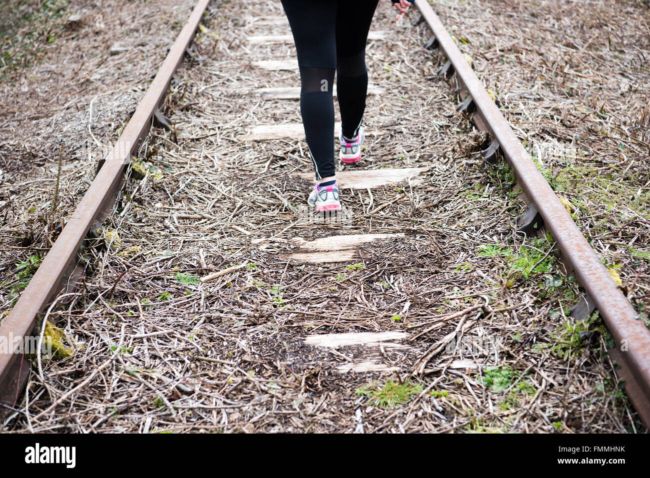 Woman's legs in black sports leggings and trainers walking away from the viewer in the middle of a disused rail track Stock Photo