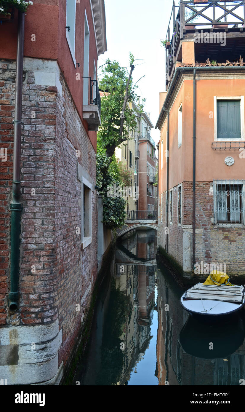 A small canal running between two old buildings in Venice. A boat is moored to one side; a bridge arches over the water. Reflections are in the water. Stock Photo