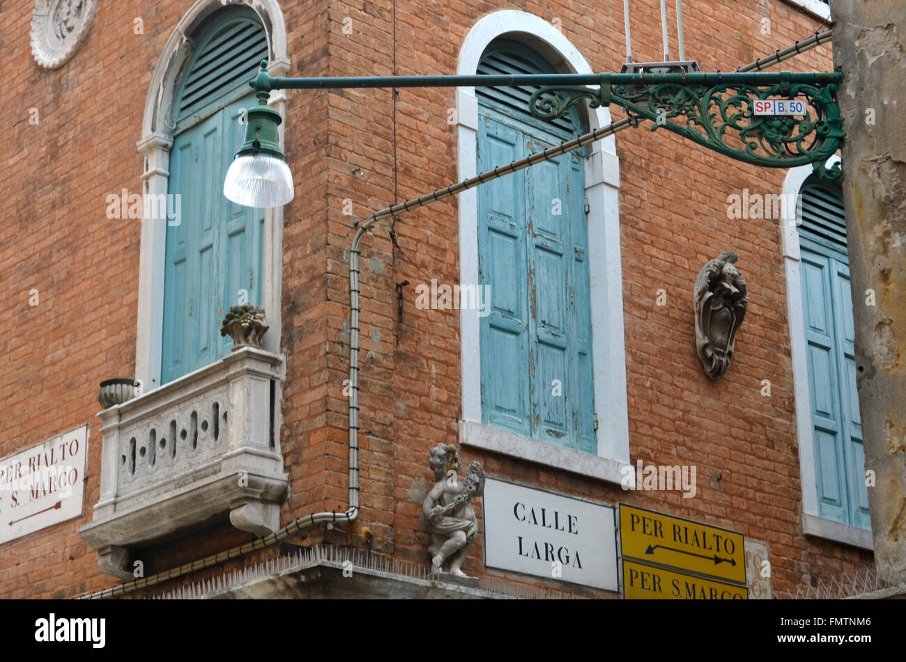 Street signs on the corner of an old Venetian building, plus a marble balcony, a stone angel and a coat of arms. The windows have green shutters. Stock Photo