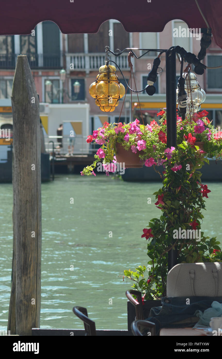 View from a restaurant across a canal in Venice. Red and pink geraniums abound, and a yellow lantern hangs from the awning. Stock Photo