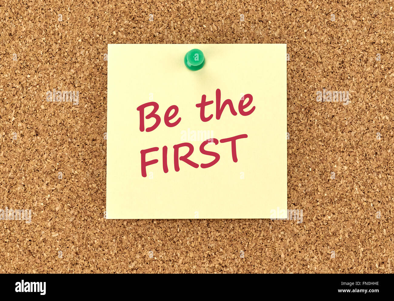 The phrase Be The First in red text on a yellow sticky note posted to a cork notice board. Stock Photo