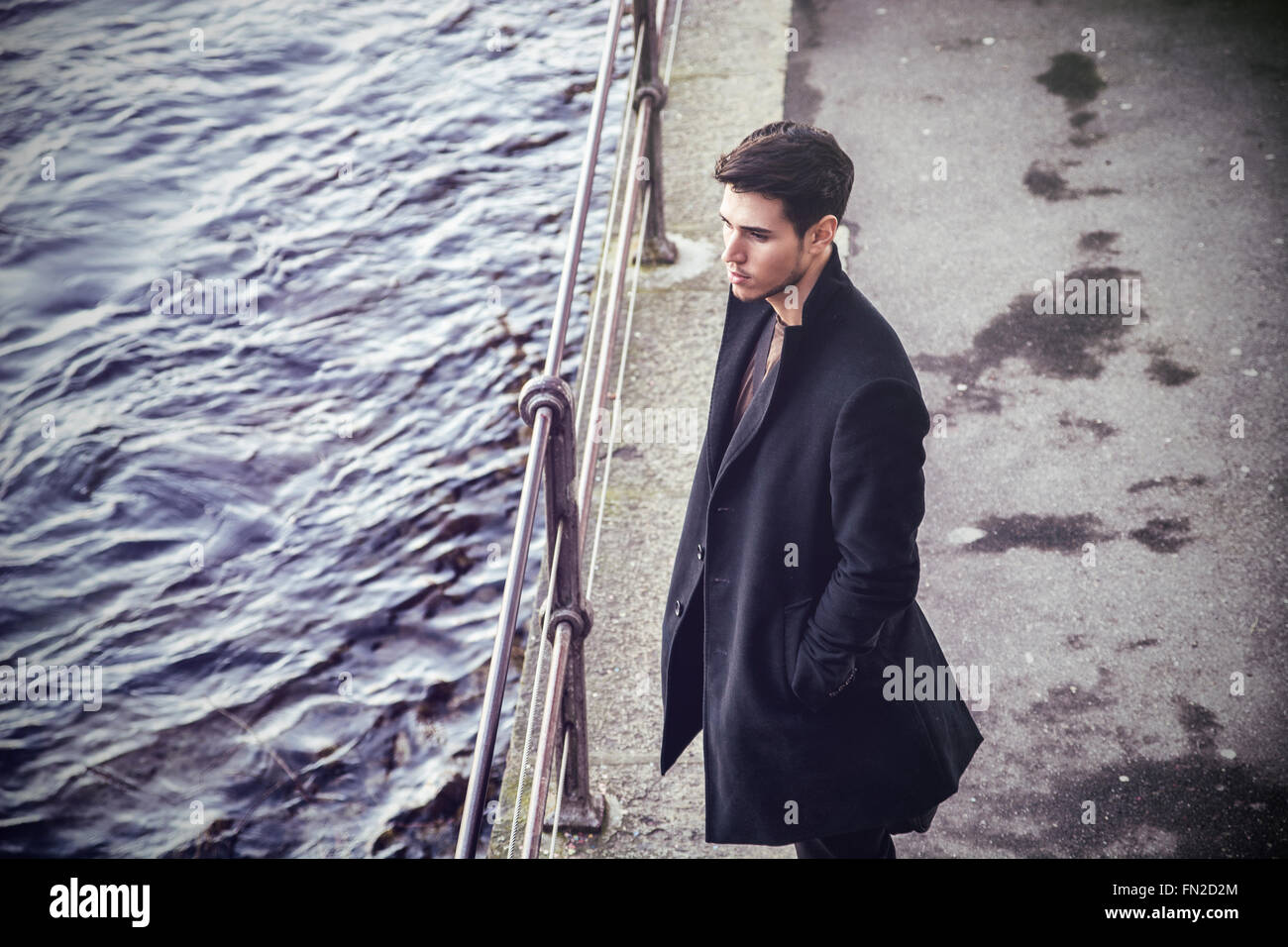Sad brunet in black clothes looking down while standing near railing outdoors. Stock Photo