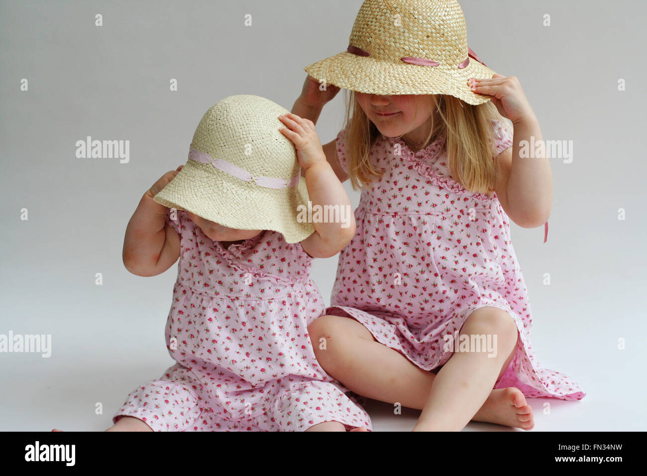 Two blonde little girls,children sitting on the floor playing, matching pink dresses and straw hat easter bonnets, easter concept, siblings ,childhood Stock Photo