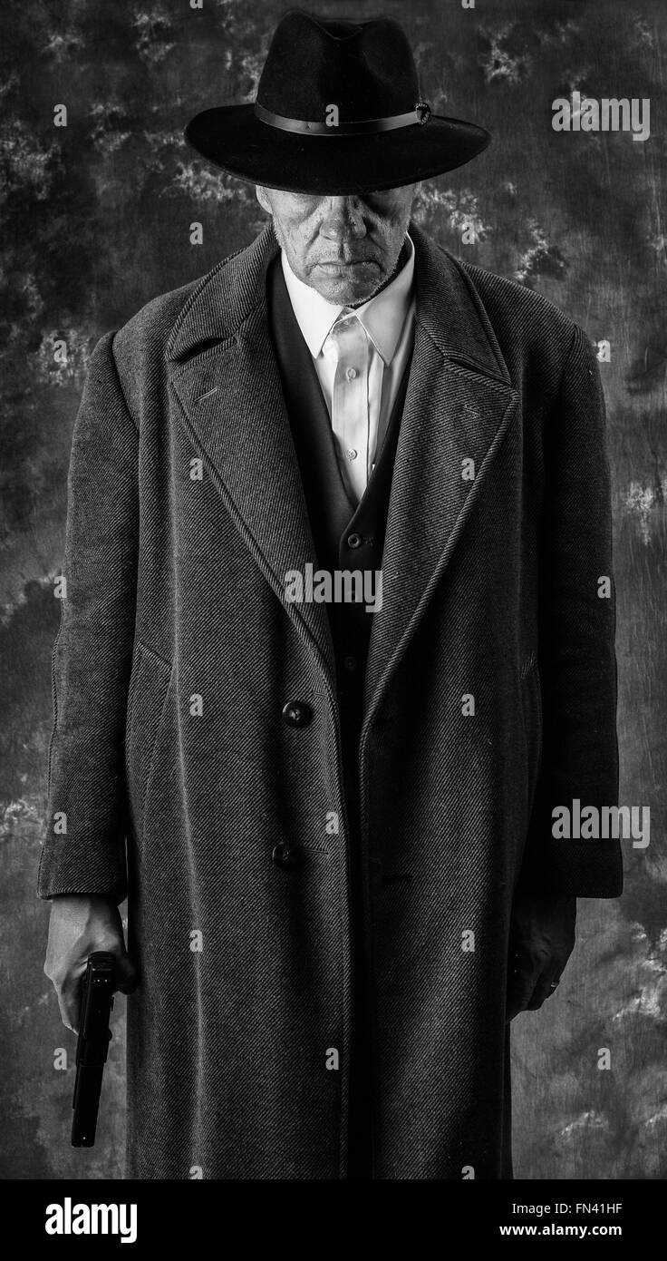 Black & white image of sinister looking man wearing overcoat and hat holding automatic pistol against portrait background Stock Photo