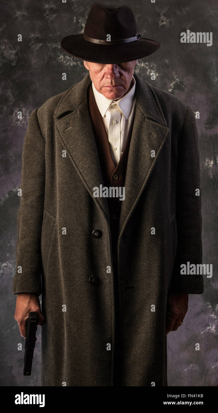 Image of sinister looking man wearing overcoat and hat holding automatic pistol to one side against portrait background Stock Photo