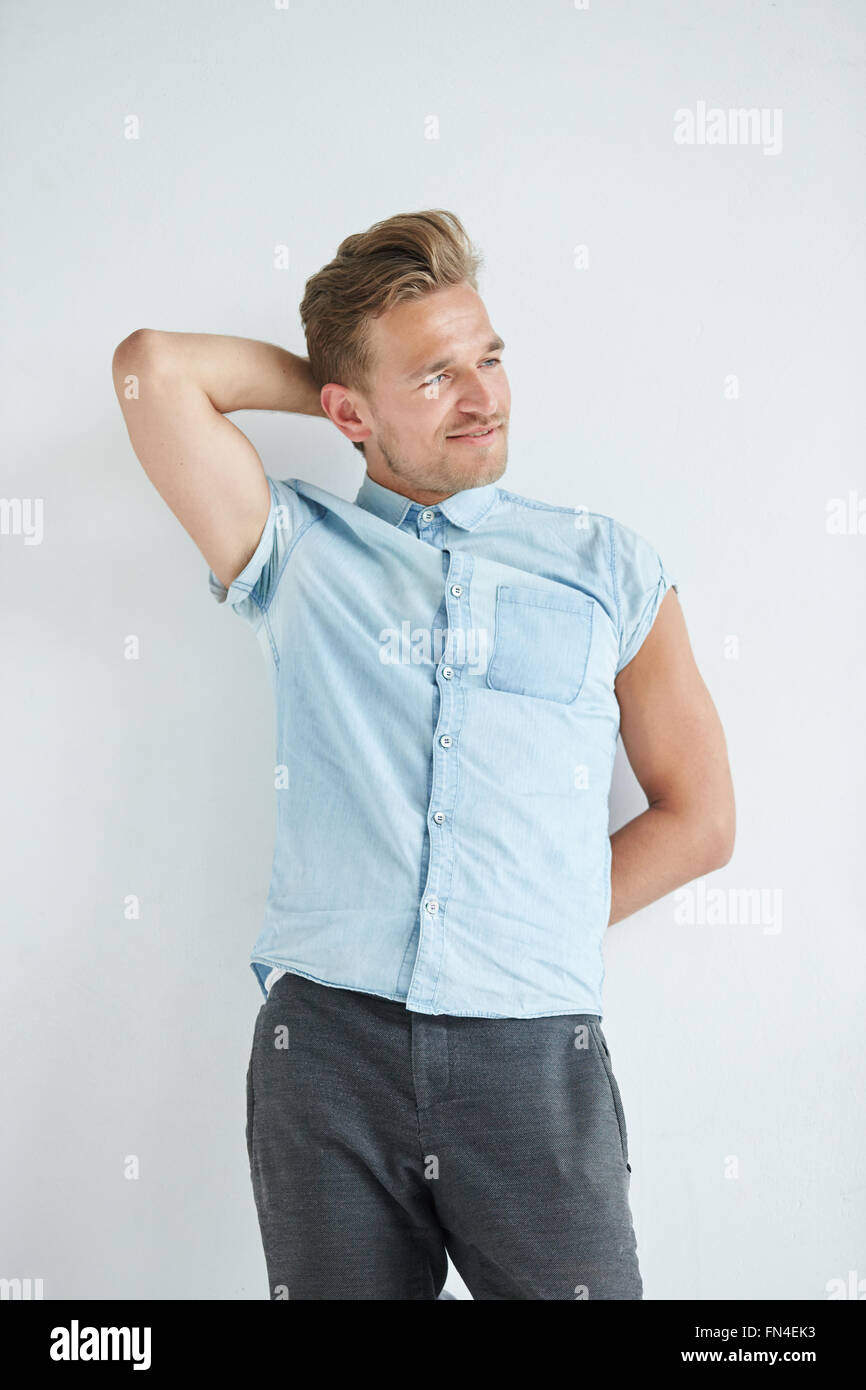 Brutal man with a small beard is leaning on the wall , wearing a blue short-sleeved shirt and gray pants , hands are behind his  Stock Photo