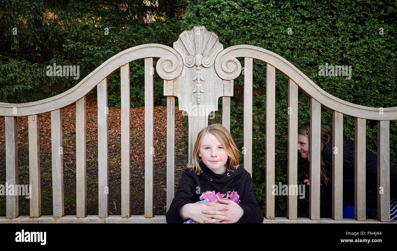 A young girl posing on a bench, her sister creeps up behind ready to scare her. Stock Photo