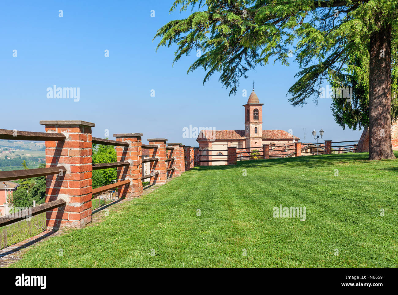 Brick fence along green lawn as small parish church on background under blue sky in Piedmont, Northern Italy. Stock Photo