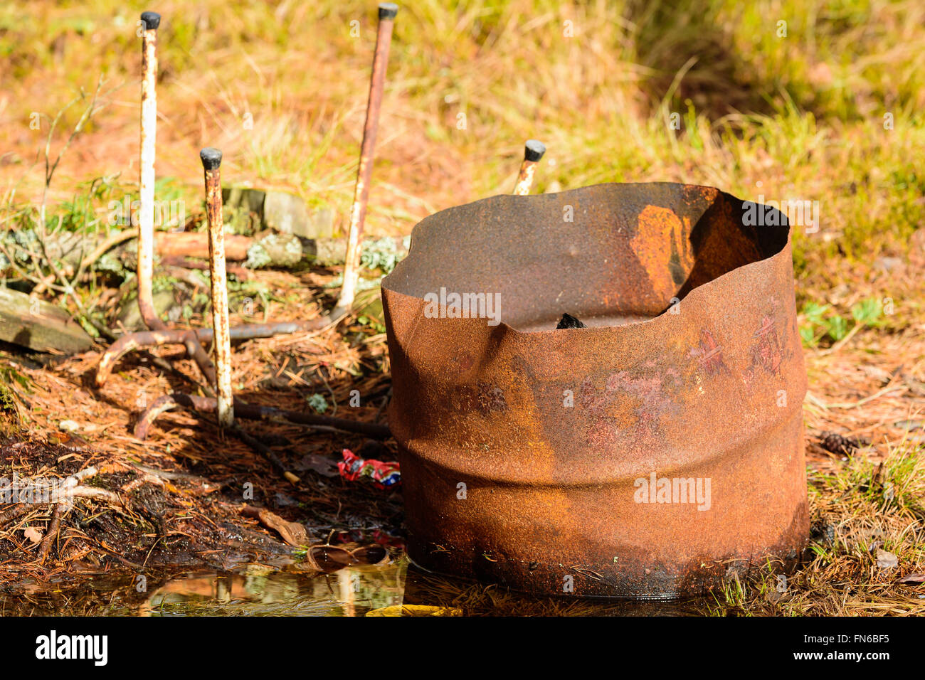 The bottom half of a rusty old barrel standing in damp and wet soil. Part of old chair in background. Stock Photo