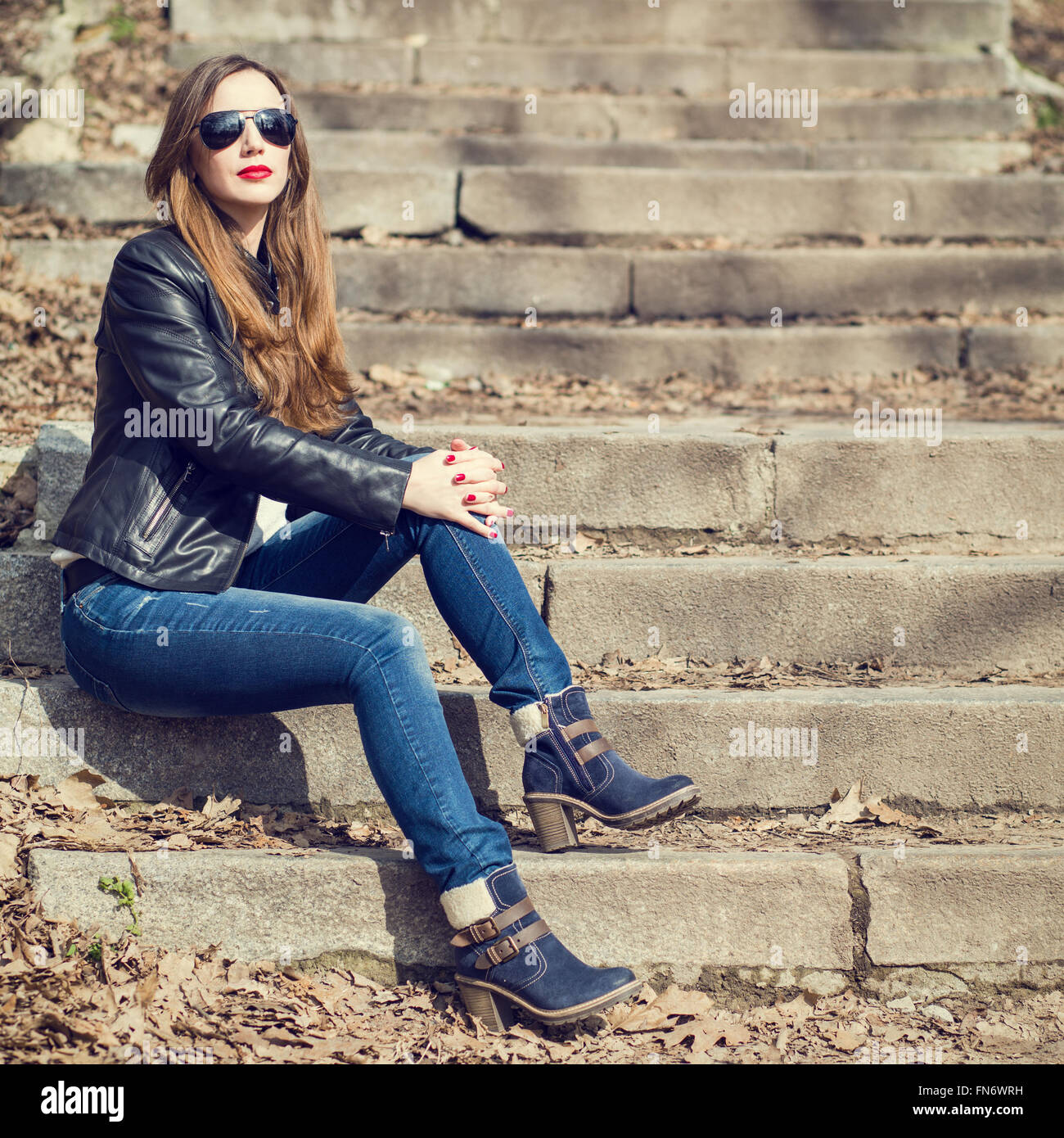 Young stylish woman in jeans and leather jacket resting on the park stairs. Attractive girl with long hair sitting on steps Stock Photo