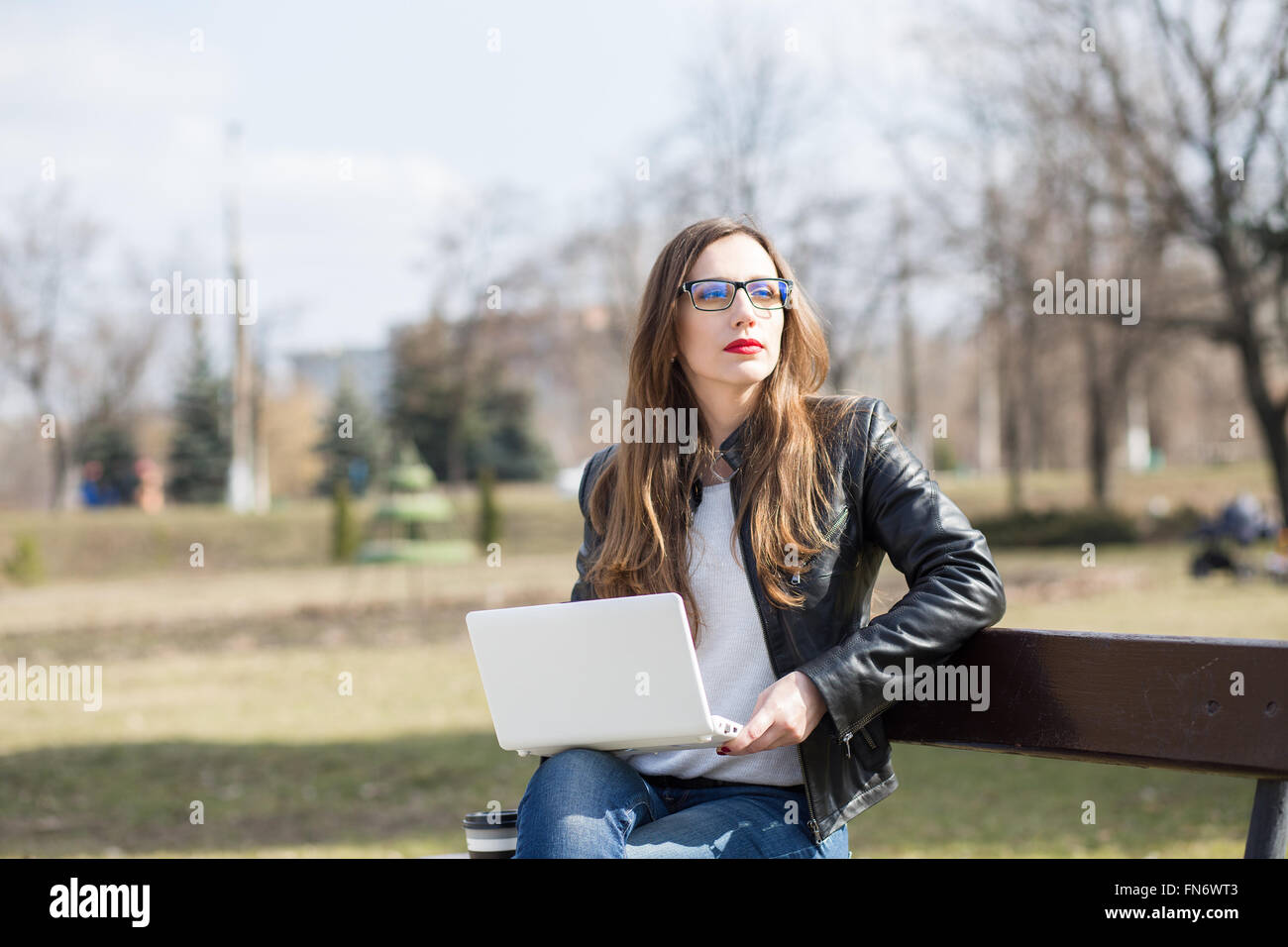 Young woman in glasses using laptop sitting on the bench in city park. Outdoor workplace background Stock Photo