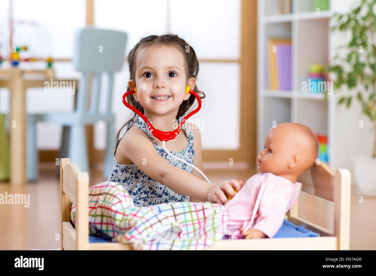 child girl playing doctor role game examining her doll using stethoscope sitting in playroom at home, school or kindergarten Stock Photo