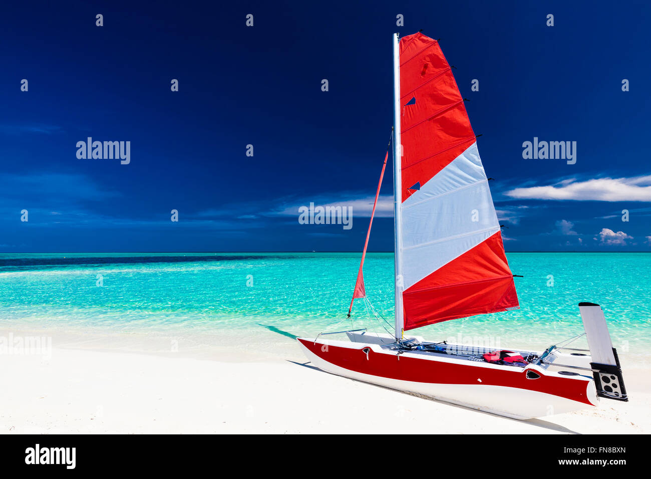Sailing boat with red sail on a beach of deserted tropical island with shallow blue water Stock Photo
