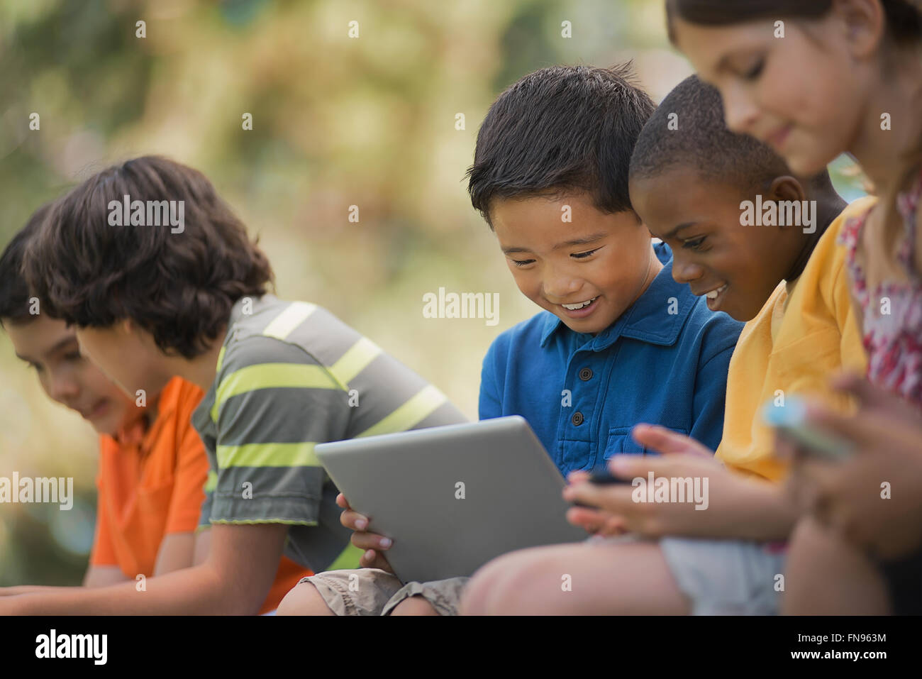 A row of children sitting outdoors in summer using tablets and handheld games. Stock Photo