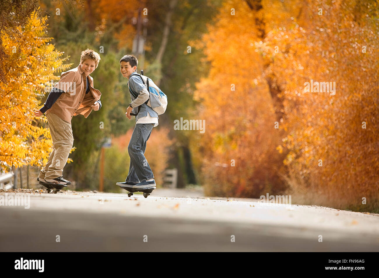 Two boys on skate boards on a roadway in woodland with vivid autumn foliage. Stock Photo