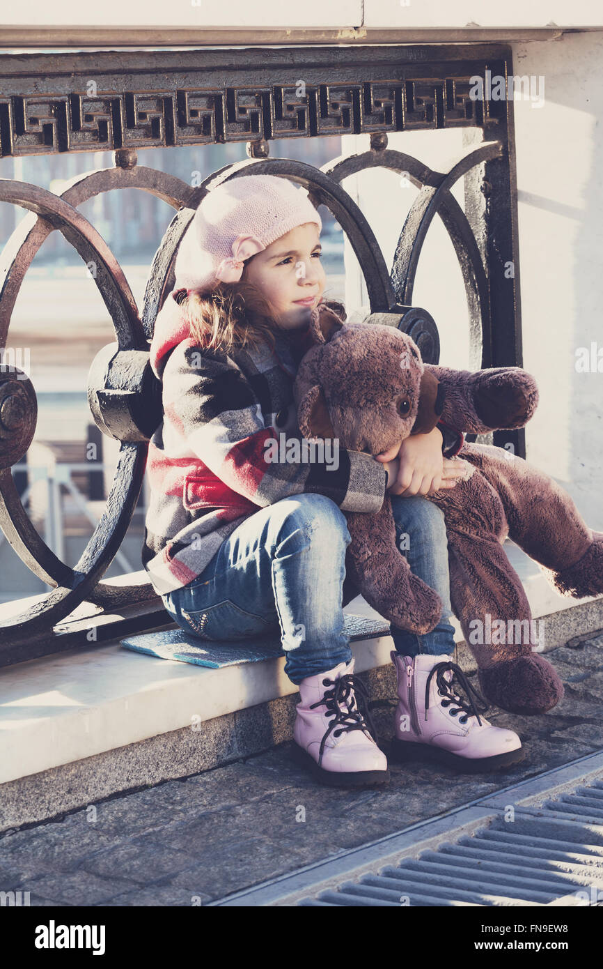 little girl in a pink beret sits at a fencing and holds a toy bear in hand Stock Photo