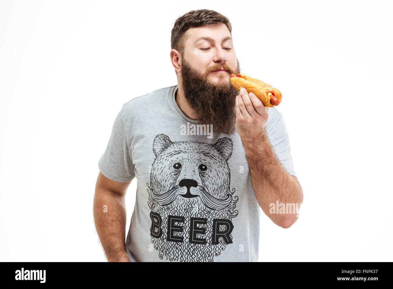 Happy bearded man standing and eating hot dog over white background Stock Photo