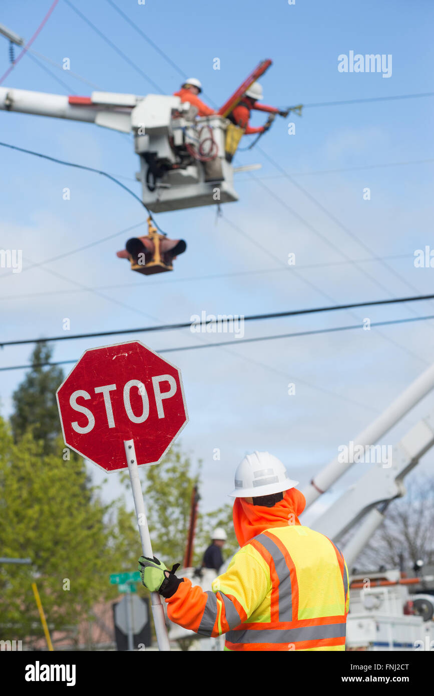 A traffic guard holds a stop sign as two power line repairmen hover in the sky, repairing an electric line. Stock Photo