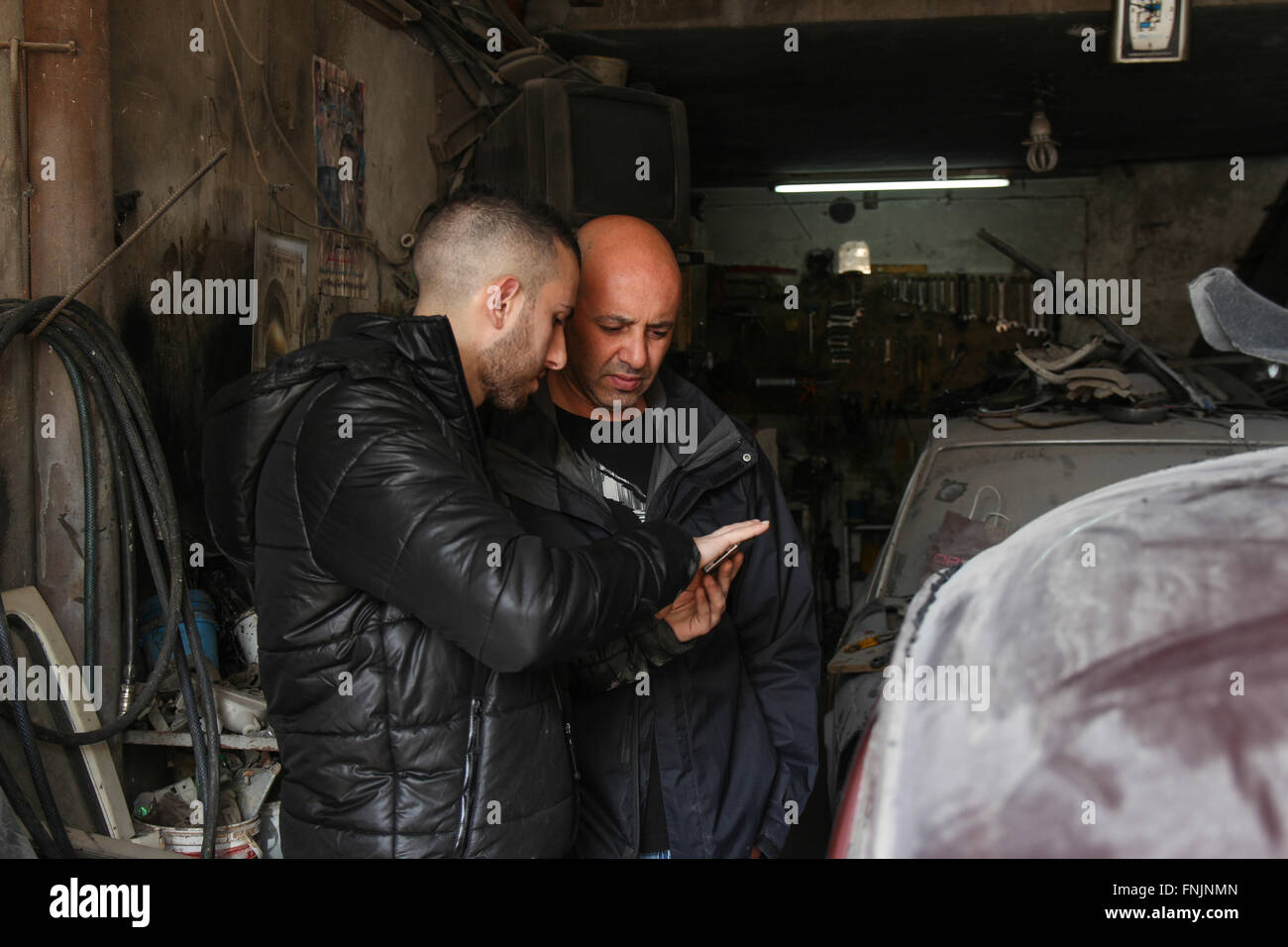 Nablus, Palestinian Territories. 11th Feb, 2016. Ali Al-Kutob (L) and Omar Zuhairi look at a smartphone as they stand next to Zuhairi's Volkswagen Beetle in a car repair shop in Nablus, Palestinian Territories, 11 February 2016. An increasing number of Palestinians are discovering the classic car for themselves. Photo: DAVID EHL/dpa/Alamy Live News Stock Photo