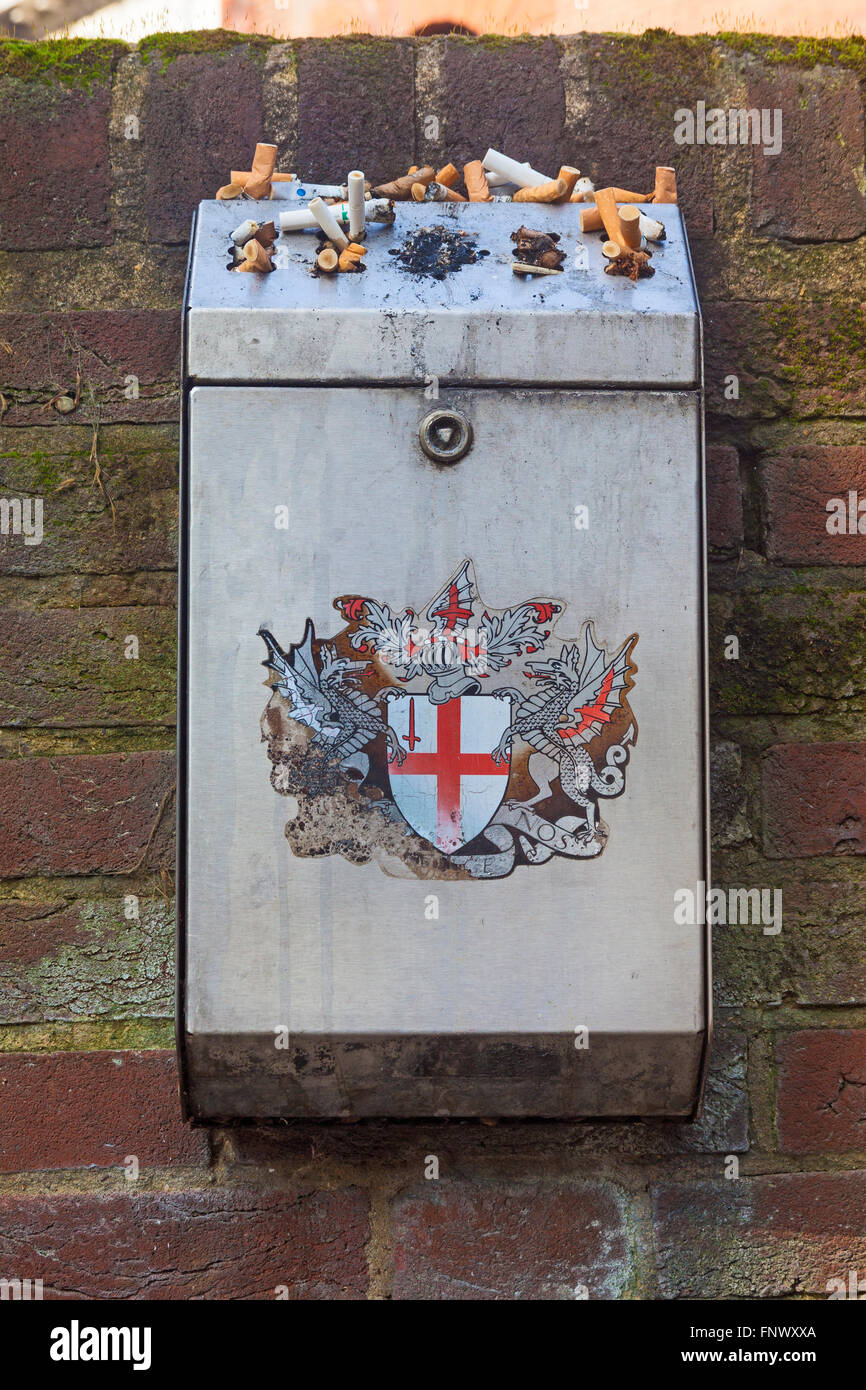 A wall-mounted receptacle for cigarette ends, complete with the much-worn City  of London coat of arms, in a City churchyard Stock Photo