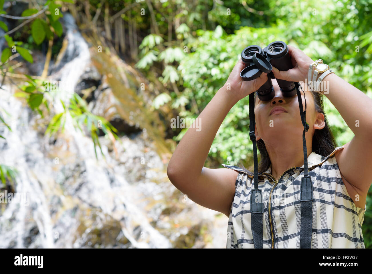 Beautiful young girl hiking is using binoculars look for birds in tropical forest near the waterfalls in Thailand Stock Photo