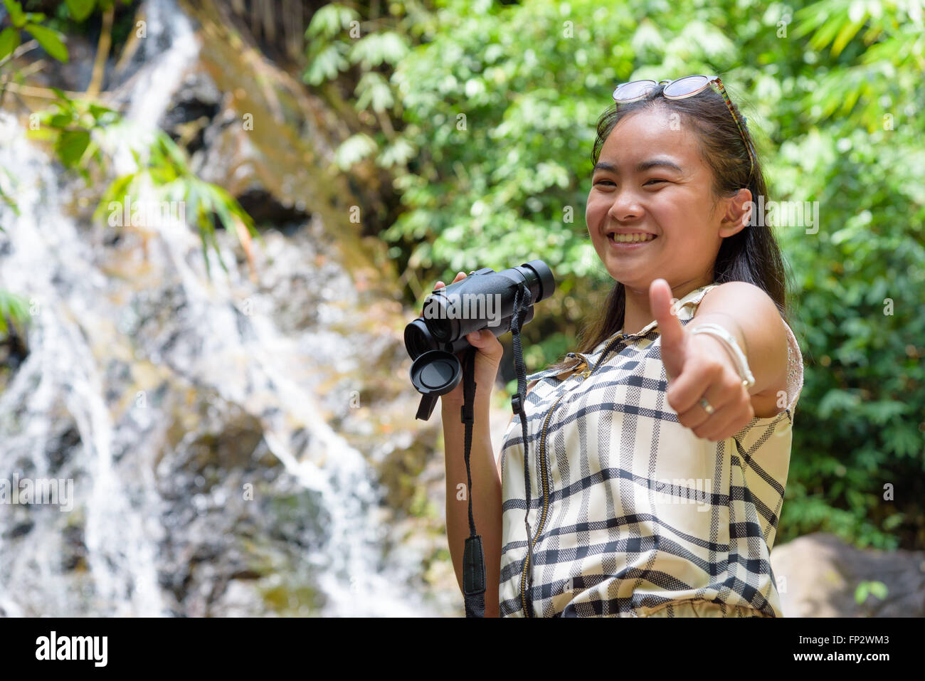 Beautiful young girl hiking is using binoculars look for birds smiled and raised a thumb to the camera in tropical forest Stock Photo