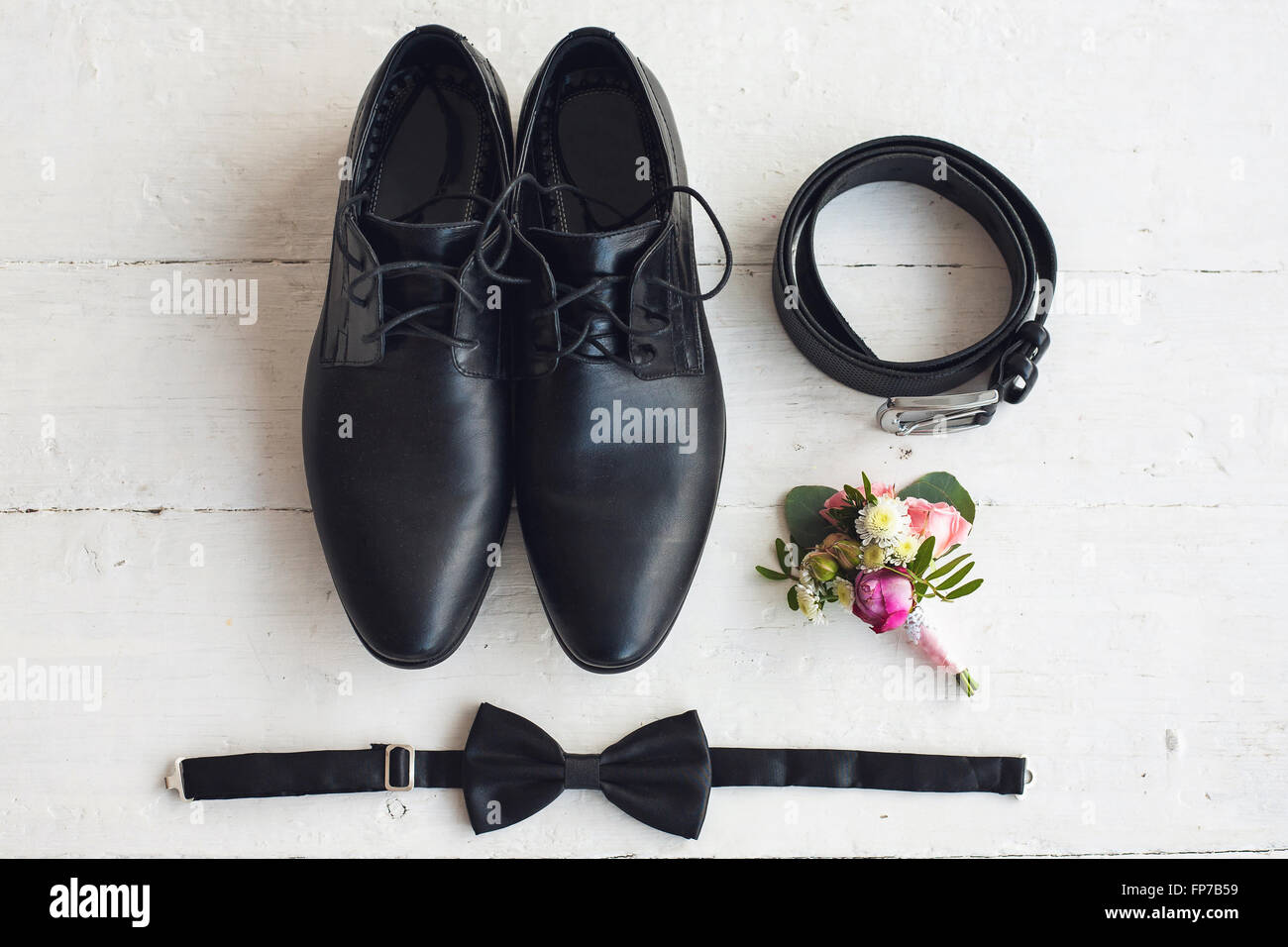 Close up of modern man accessories. Black bowtie, leather shoes, belt and flower boutonniere on white wood rustic background. Se Stock Photo