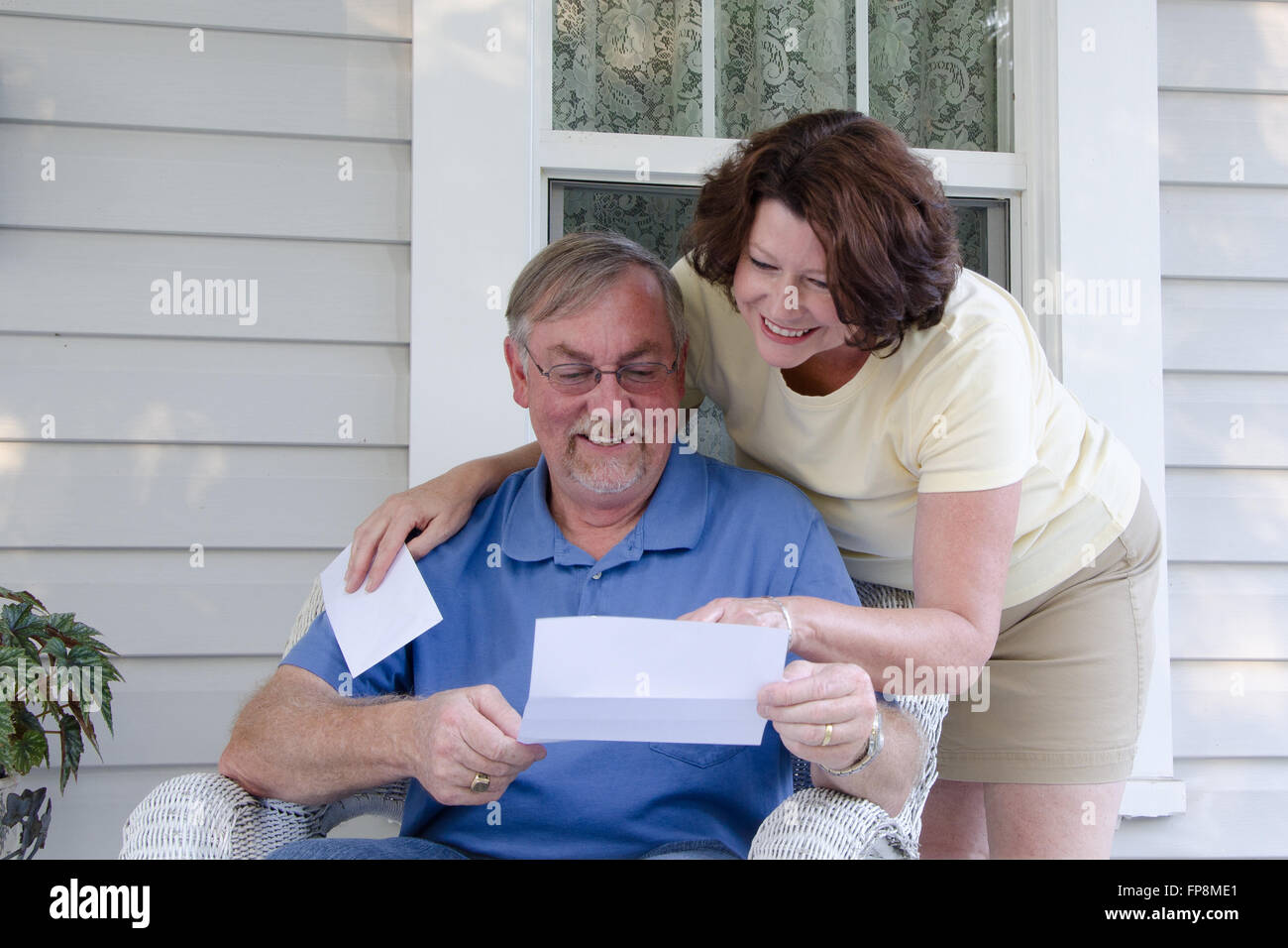 A middle aged couple reads a letter on the porch of their vintage house, enjoying reading good news together. Stock Photo