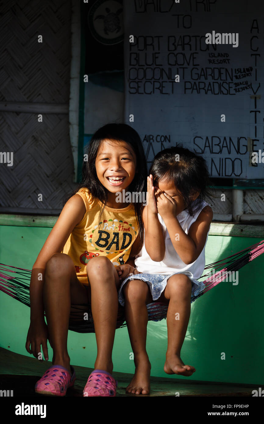 Two girls sitting in the sun in a hammock strung up on the wall in front of a local store smiling, laughing, little one hiding Stock Photo