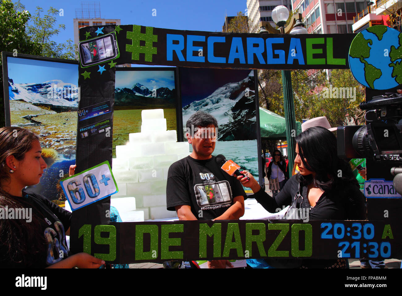 La Paz, Bolivia, 18th March 2016. An activist talks about Earth Hour (which takes place tomorrow) to a local TV press reporter at an event organised by the La Paz City Government. Behind him blocks of ice melt in front of photos of Bolivia's mountains, a display to highlight global warming (glaciers in Bolivia and the Andes are some of the fastest receding on the planet). The slogan reads 'Recarga el Planeta' / 'Recharge the Planet'. Credit: James Brunker / Alamy Live News Stock Photo