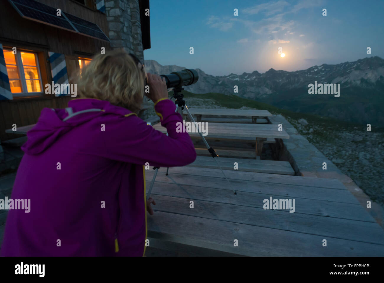 A woman looks at the full moon over Prättigau mountains with a telescope on the Carschina mountain hut in the Swiss Alps,Grisons Stock Photo