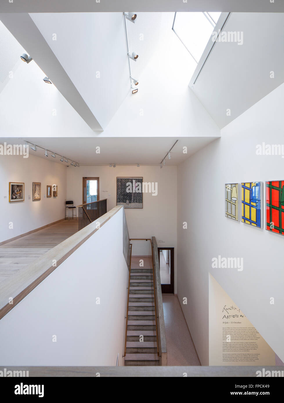 A stairway in an art gallery with art on the walls, light coming through roof lights, against a