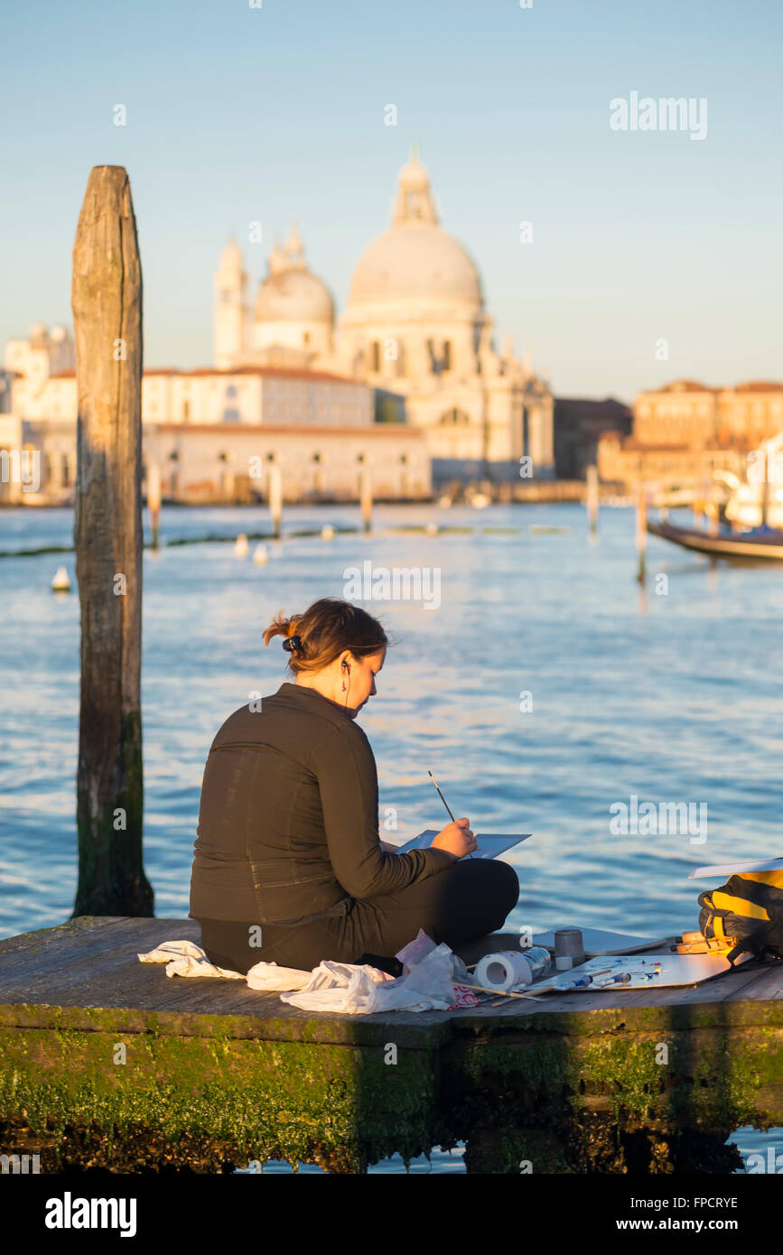 Woman sitting on a jetty near the St. Mark's Square at sunrise painting the cityscape of Venice at the Grand Canal Stock Photo