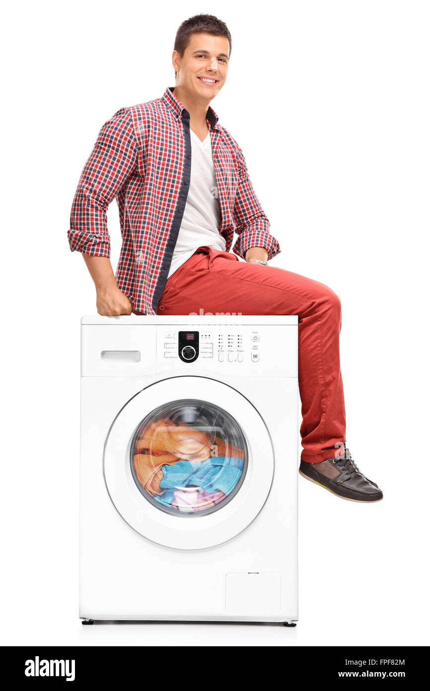 Vertical shot of a young man waiting for the laundry seated on a washing machine isolated on white background Stock Photo
