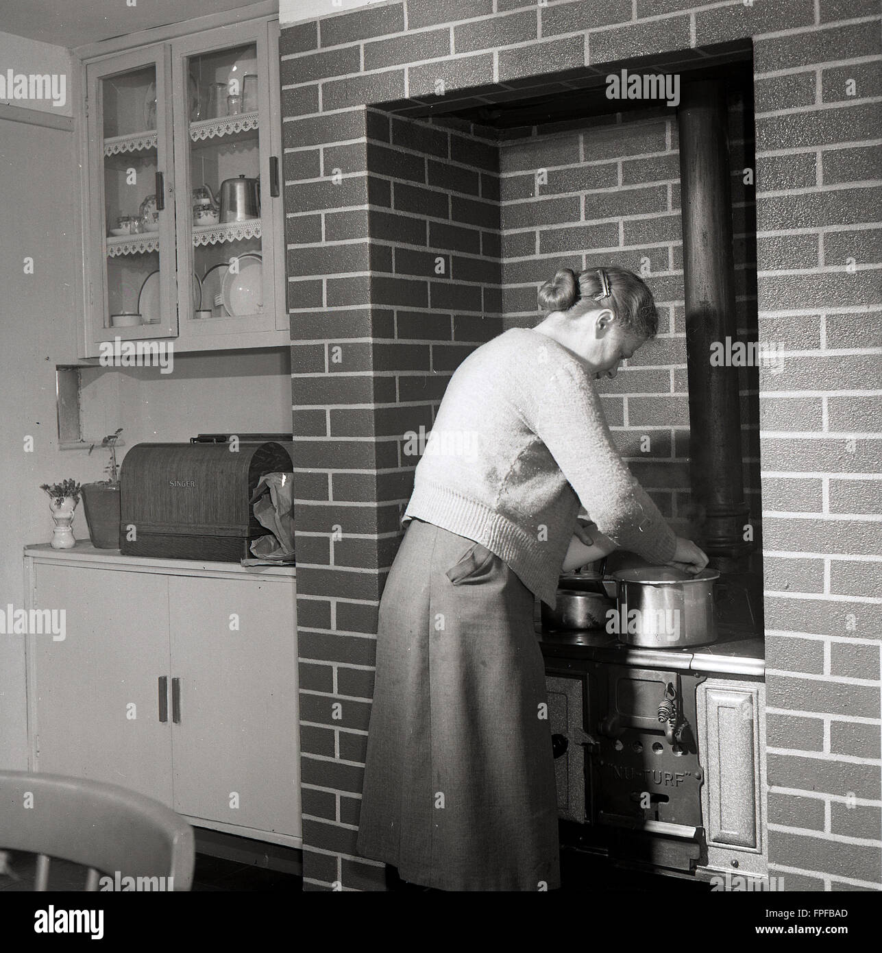 1950s historical, a lady at her stove or range, built into a brick chmney fireplace surround, checking on a metal pot, Ireland. When stoves replaced open fires for heating a property, models were developed that could also be used for cooking and these were referred to as ranges, as many had more than one oven and two cooking spots on top, giving a 'range' of places to cook on. Stock Photo