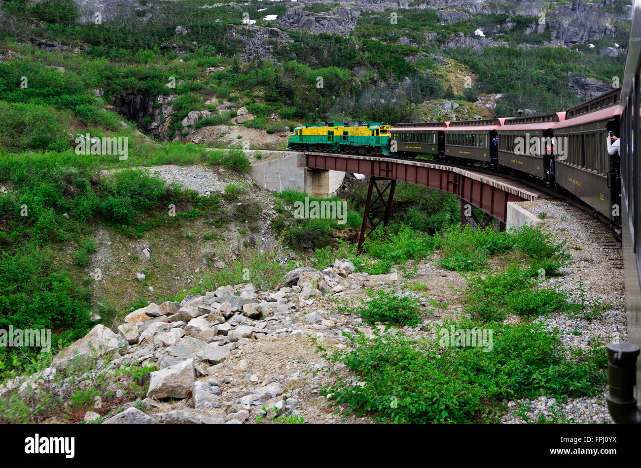 White Pass Railroad electric engine pulls train passenger cars over trestle bridge towards tunnel in the rocks. Stock Photo