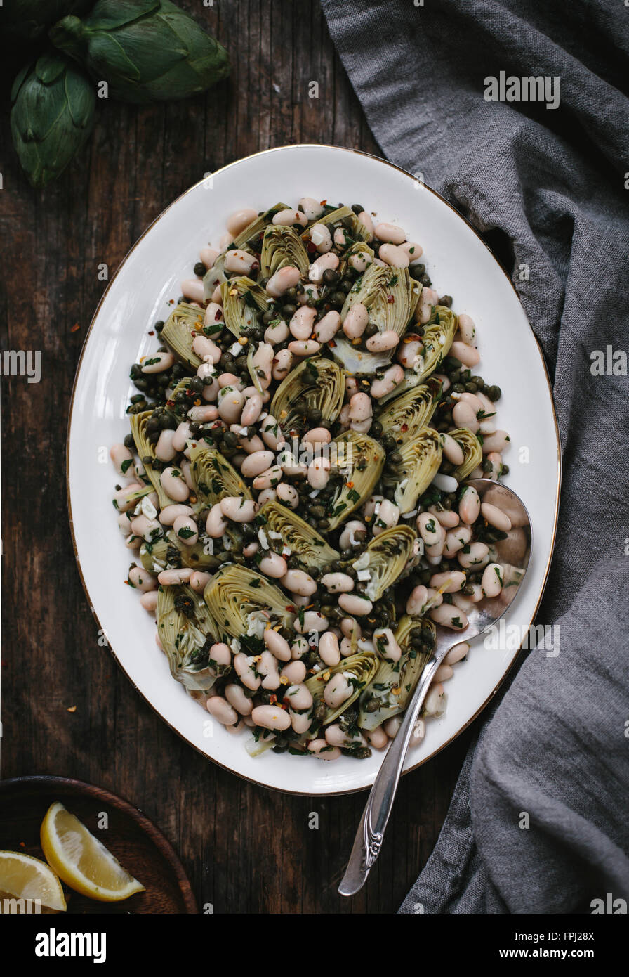 An oval plate filled with braised baby artichoke salad with white beans and capers is photographed from the top view. Stock Photo