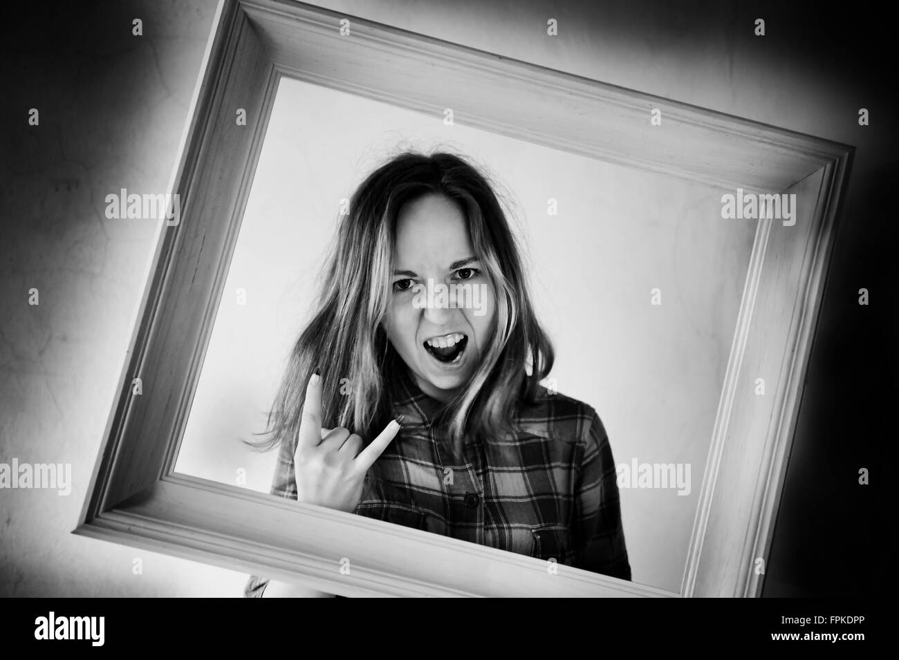 Crazy woman posing in frame, adult girl with funny mad face and rock and roll hand horns gesture, photographed in a wooden frame Stock Photo
