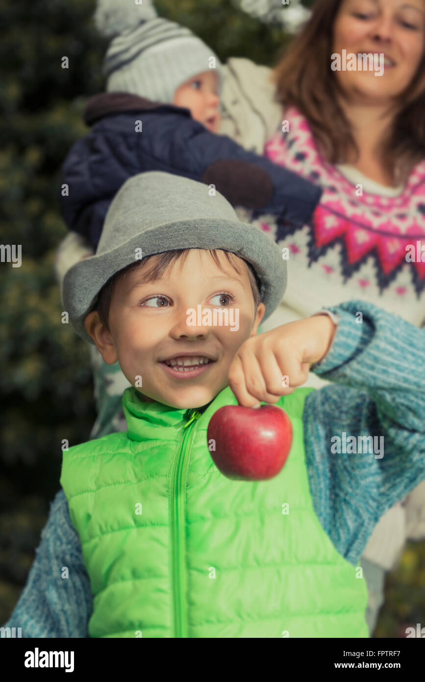 Little boy showing red apple and standing mother with baby in the background, Bavaria, Germany Stock Photo