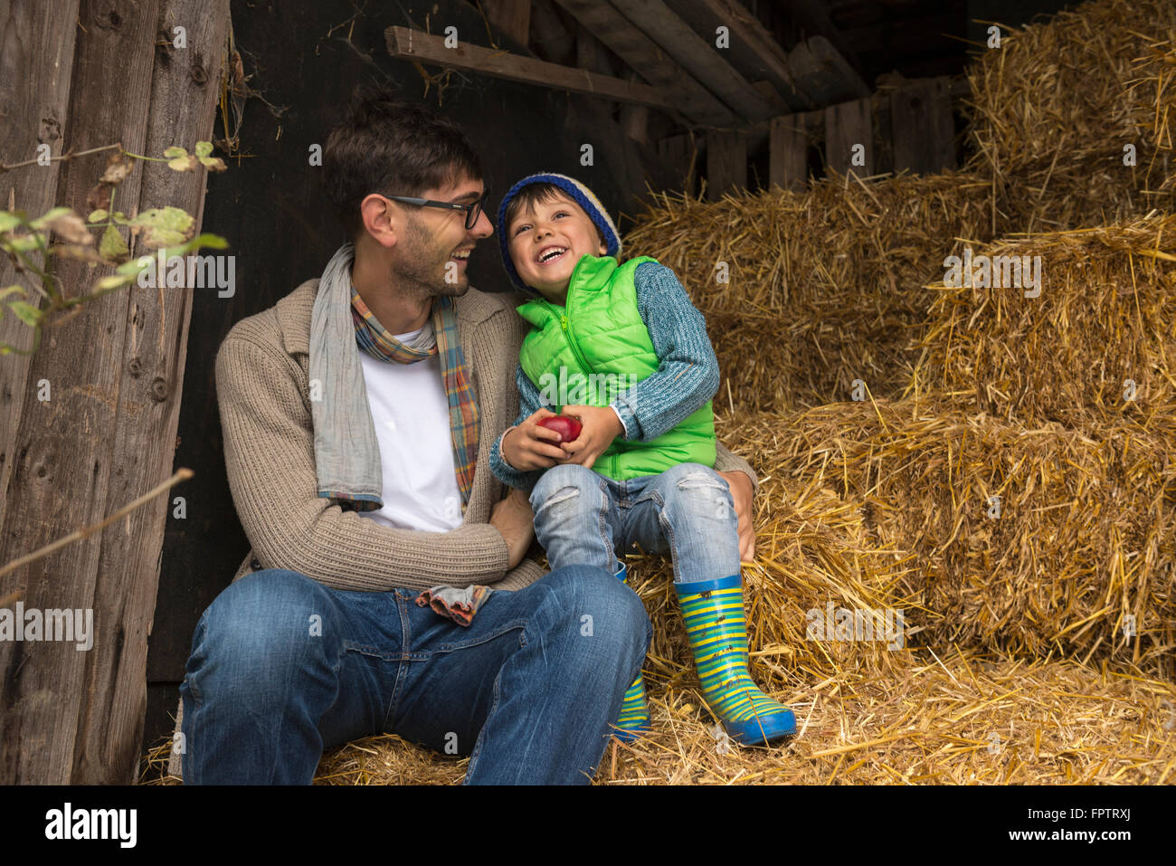 Father and son sitting on straw in the stable and laughing, Bavaria, Germany Stock Photo
