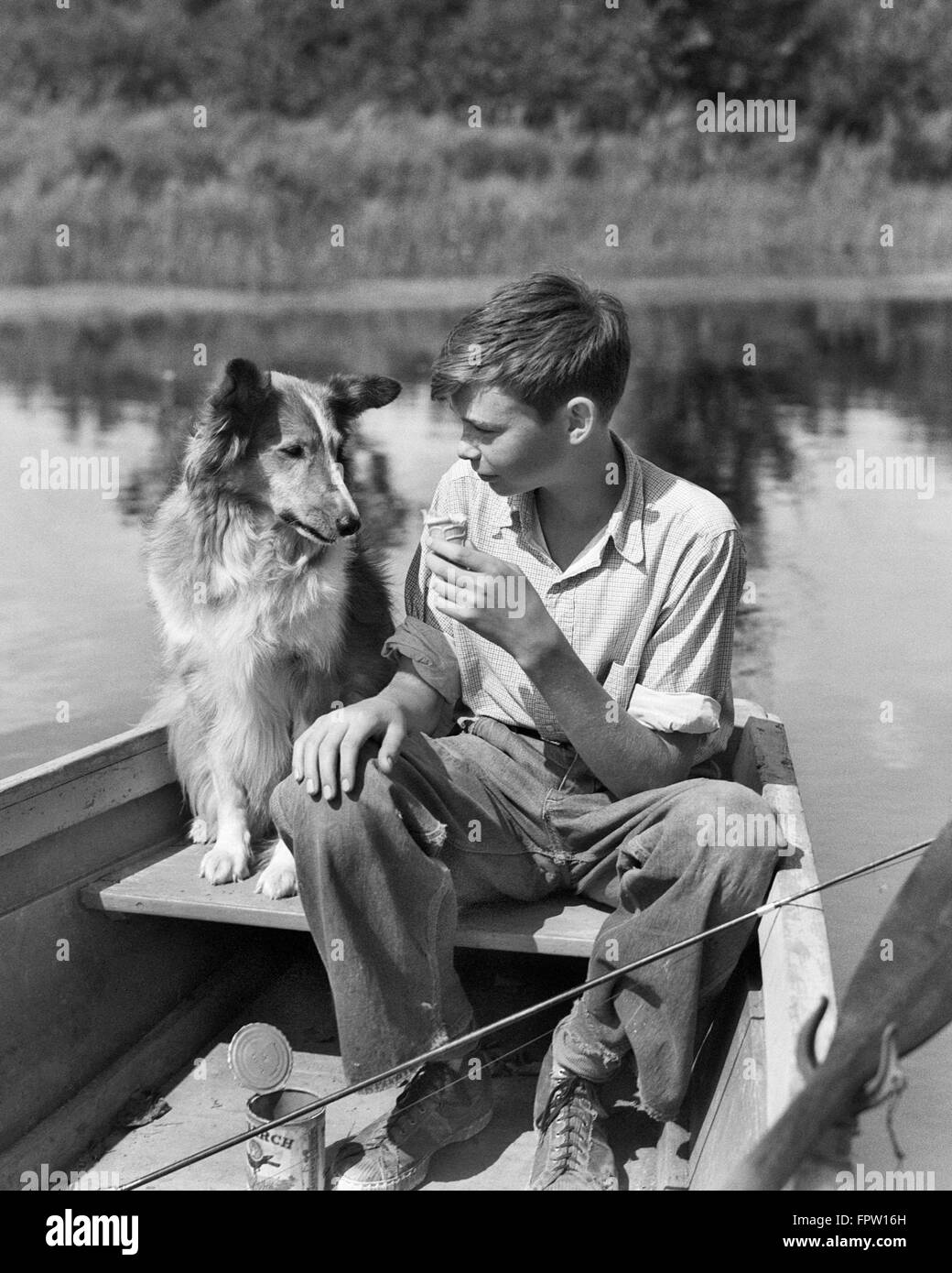 1930s BOY AND COLLIE DOG TOGETHER IN ROWBOAT WITH FISHING POLE CAN OF WORMS ON POND EATING SHARING ICE CREAM CONE Stock Photo