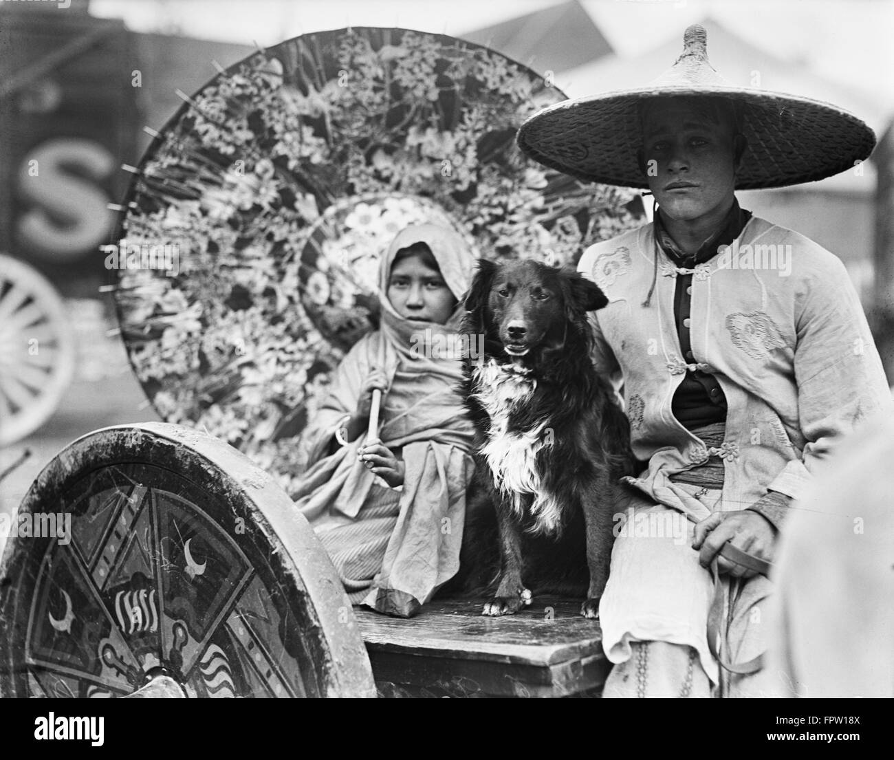 1930s PORTRAIT OF MAN AND GIRL CIRCUS PERFORMERS WITH DOG WEARING TRADITIONAL ASIAN CLOTHING GIRL HOLDING DECORATIVE UMBRELLA Stock Photo