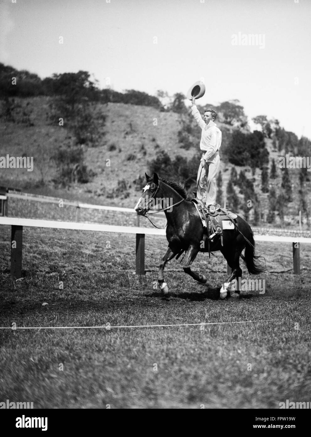 1930s JIM ROGERS SON OF ACTOR WILL ROGERS PERFORMING STUNT TRICK STANDING ON GALLOPING HORSE BACK WAVING COWBOY HAT Stock Photo