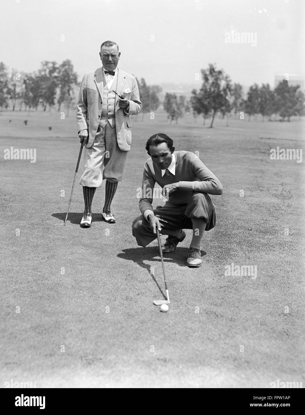 1920s TWO MEN WEARING PLUS FOURS PLAYING GOLF ONE MAN SMOKING PIPE OTHER MAN KNEELING BY GOLF BALL LINING UP PUTT Stock Photo