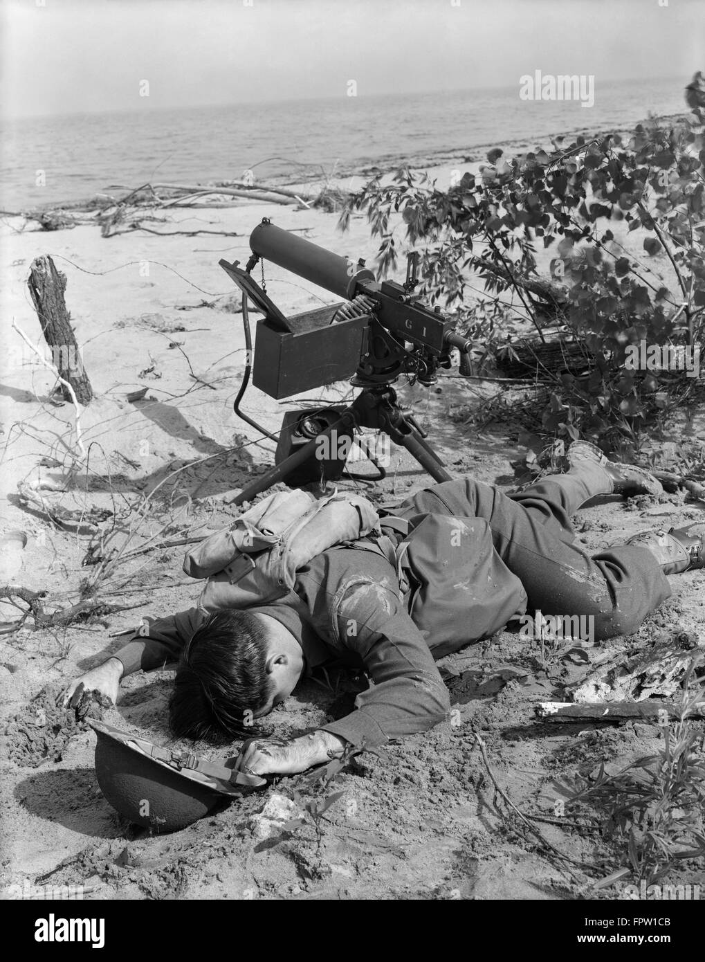 1940s WORLD WAR II INJURED DEAD SOLDIER LYING FACE DOWN ON OCEAN BEACH NEXT TO MACHINE GUN Stock Photo