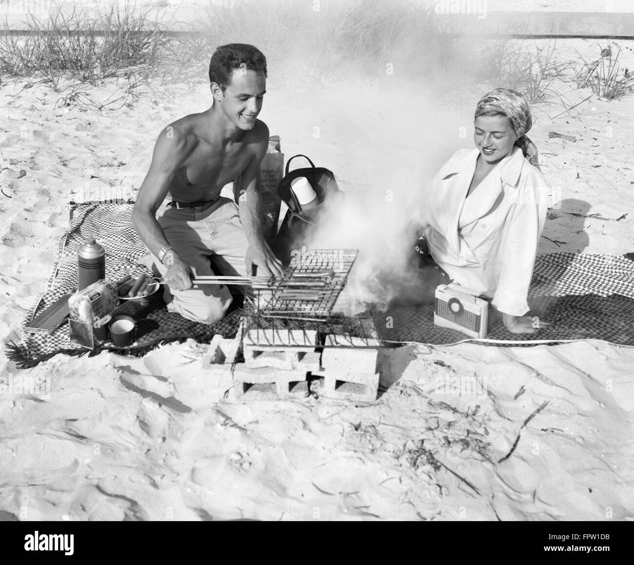 1940s COUPLE HAVING PICNIC ON BEACH MAN GRILLING HOT DOGS Stock Photo