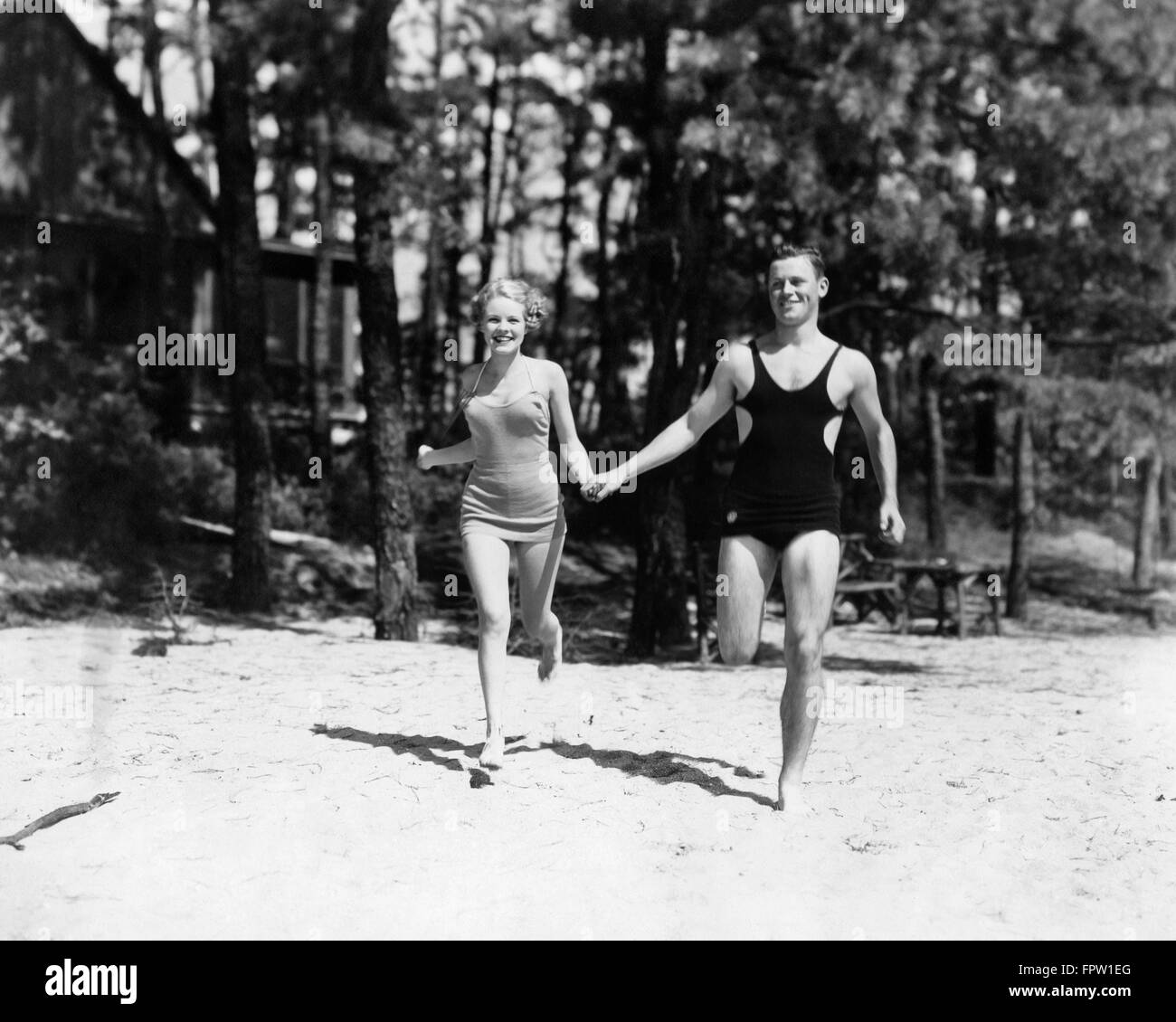 1930s COUPLE WEARING BATHING SUITS HOLDING HANDS RUNNING ON BEACH Stock Photo
